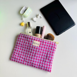 May include: A pink and white gingham print cosmetic bag with a gold zipper, open to reveal makeup items. A yellow star charm hangs from a light green and yellow patterned AirPod case. A black tablet and white earbuds are also visible.