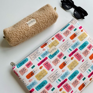 Puede incluir: Un estuche beige y difuso y una bolsa blanca con cremallera con un patrón de etiquetas coloridas. Las etiquetas tienen frases como "Perfect", "Smart" y "Awesome". También hay gafas de sol negras en la imagen.