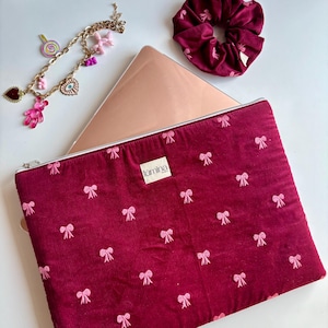Könnte beinhalten: Eine bordeauxfarbene Laptoptasche mit rosa Schleifenmuster, ein passendes Scrunchie und ein Charm-Armband mit verschiedenen Anhängern. Die Laptoptasche hat einen Reißverschluss und ein Etikett mit der Aufschrift "lumina".