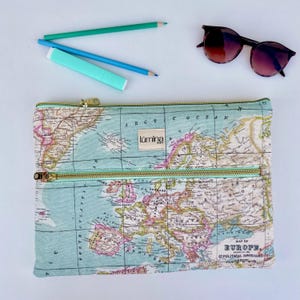 Puede incluir: Una bolsa con cremallera azul claro con estampado de mapa y cremalleras doradas. La bolsa tiene un ribete verde claro y una pequeña etiqueta que dice "lumina". También hay dos lápices y un par de gafas de sol en la imagen.