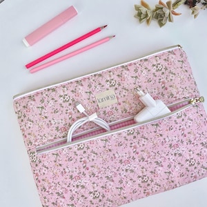 May include: A rectangular fabric pouch featuring a pink floral pattern with a zippered front pocket. The pouch has a small beige label with the text "lumina" in black. Inside the open front pocket are a white charging cable and a white power adapter.