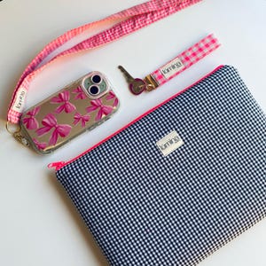 Könnte beinhalten: Eine Sammlung von Accessoires, darunter eine Handyhülle, ein Schlüsselband, eine Handschlaufe und eine Tasche. Die Handyhülle ist transparent mit rosa Schleifenmotiven. Das Schlüsselband und die Handschlaufe sind rosa und weiß kariert. Die Tasche ist schwarz-weiß kariert mit einem rosa Reißverschluss.