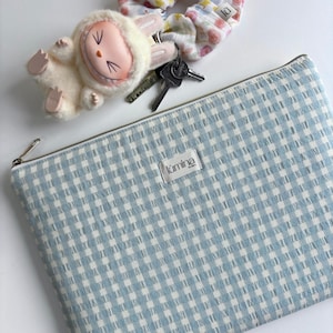 May include: A blue and white gingham zippered pouch with the word "kumina" printed on a white label. The pouch is lying on a white surface.