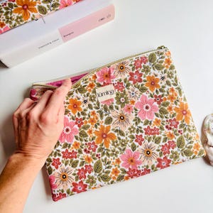 Può includere: Una pochette con cerniera a fantasia floreale con cerniera dorata. La pochette presenta uno sfondo bianco con un motivo ripetuto di fiori rosa, arancioni e rossi con foglie verdi. È visibile il nome del marchio "lúmina".