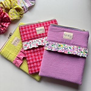 Può includere: Tre pochette con cerniera gialle, rosse e viola, ciascuna con un motivo a quadretti. Le pochette hanno un volant decorativo sul fondo con motivi floreali o di ciliegie. Ogni pochette ha una piccola etichetta con la scritta "lúmina".