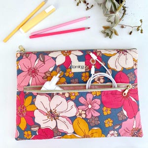 Peut inclure: Une pochette pour ordinateur portable à motifs floraux avec de grandes fleurs roses, jaunes et blanches sur fond bleu. La pochette a deux compartiments zippés, l'un contenant un chargeur blanc et l'autre un câble de chargement blanc. Des crayons et une gomme sont visibles.