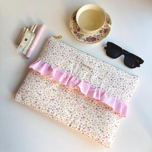 Può includere: Una pochette con stampa floreale con volant rosa a quadretti, cerniera dorata e il marchio "lumina". Sono visibili anche una tazza e un piattino, occhiali da sole e flaconi di profumo.