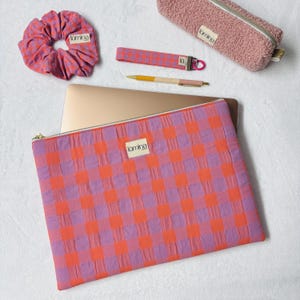 Può includere: Una custodia per laptop con motivo a quadri viola e arancione, un elastico per capelli abbinato, un portachiavi da polso, un astuccio rosa in peluche e una penna. Ogni articolo presenta una piccola etichetta bianca con la scritta Lumina.