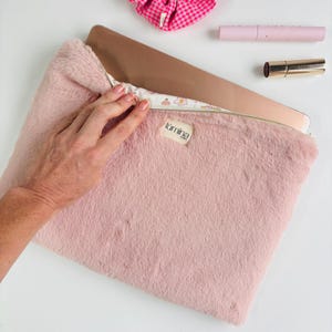 Peut inclure: Une housse d'ordinateur portable en fausse fourrure rose clair avec une fermeture éclair dorée et une petite étiquette "lumina". La housse est ouverte, révélant un ordinateur portable. Un chouchou rose, du mascara et du rouge à lèvres sont également visibles.