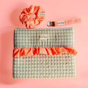 Pode incluir: Um conjunto de acessórios, incluindo uma scrunchie, um porta-chaves e uma bolsa com zíper. Os itens apresentam um padrão xadrez vichy em tons de verde e branco, com babados e detalhes em laranja brilhante. A marca "kūmina" é visível.