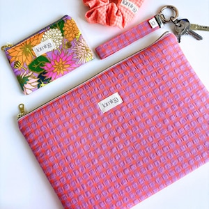Peut inclure: Une pochette zippée à carreaux rose et violet avec une fermeture éclair dorée et le mot "lumina" imprimé sur le devant. La pochette est en tissu texturé et repose sur une surface blanche.