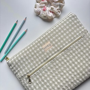 Peut inclure: Une trousse à crayons à carreaux vert clair et blanc avec une fermeture éclair dorée, l'étiquette indique "lūmina". Trois crayons dans les tons de vert et de bleu sont à côté de la trousse. Une chouchou floral avec une petite étiquette est également dans l'image.