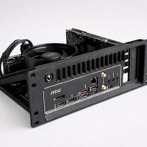 May include: A black MSI computer motherboard mounted in a black frame with a circular cooling fan, a power button, and a rear I/O panel featuring HDMI, DisplayPort, USB, Ethernet, and gold-plated antenna connectors for wireless connectivity.