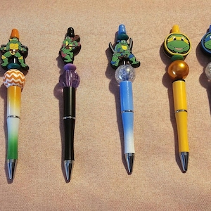 May include: A selection of seven decorative pens with Teenage Mutant Ninja Turtle character toppers. Each pen has a unique colour scheme, including white, green, blue, yellow, and red, with silver accents and a metal tip. The toppers depict the turtles.