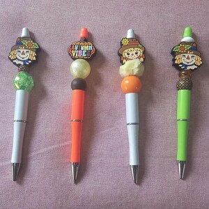 以下が含まれることがあります： 秋をテーマにした装飾的なペン6本セット。各ペンは、黄色、白、オレンジ、緑、紫など、異なる色のバレルを備えています。トップには、かかしやカボチャのデザイン、装飾的なビーズが含まれています。