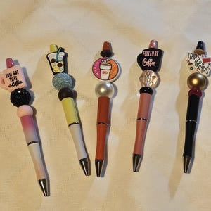 May include: Five decorative pens with various toppers. One pen has a pink mug topper with the text "YOU GOT THIS - Coffee". Other toppers include a bubble tea, a Dunkin' Donuts logo, a coffee cup with the text "FUELED BY Coffee", and a travel-themed design.