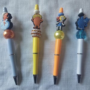 Könnte beinhalten: Vier dekorative Stifte mit Figuren als Aufsätze. Die Stifte haben verschiedenfarbige Schäfte, darunter Hellblau, Gelb und Orange. Jeder Stift hat ein einzigartiges Charakterdesign, das einen spielerischen Touch verleiht.