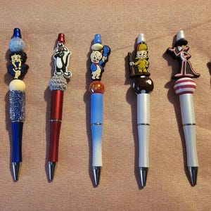 Könnte beinhalten: Sammlung dekorativer Stifte mit Aufsätzen von Zeichentrickfiguren. Die Stifte haben rote, blaue und weiße Schäfte mit silbernen Akzenten. Jeder Stift ist mit einer anderen Figur versehen, darunter ein gelber Vogel, eine blaue Figur und eine schwarz-weiße Katze.