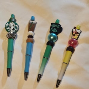 Puede incluir: Cuatro bolígrafos coloridos con diferentes encantos. Los encantos incluyen un logotipo de Starbucks verde, una taza de café helado marrón, una taza de café negra con el texto "Fueled by Coffee" y una taza de té helado amarilla.