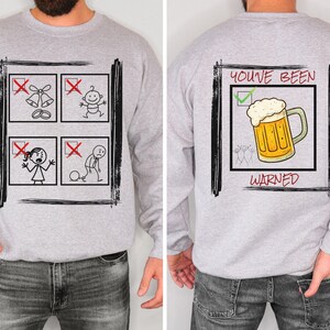 Może przedstawiać: Jasnoszara bluza z zabawnym wzorem. Przód przedstawia cztery kwadraty z przekreślonymi ilustracjami ludzików, w tym dzwony weselne, dziecko, kobietę i mężczyznę z kulą u nogi. Tył przedstawia kufel piwa z napisem "YOU'VE BEEN WARNED".