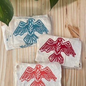 May include: A collection of rectangular fabric patches featuring a block-printed bird design. The patches are made of off-white canvas with frayed edges. The bird motif is printed in teal and red ink. Some patches feature the initials S.A.S.