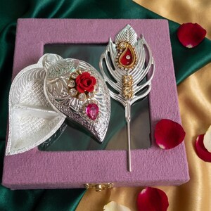 May include: A silver-toned leaf-shaped container with a red rose and pink crystal accent, paired with a matching silver-toned feather-shaped decorative stick featuring a red paisley-style inlay, all resting on a square purple textured box.