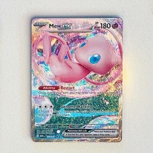 May include: A holographic Mew ex Pokémon trading card. The card features a pink Mew character with blue eyes, surrounded by a shimmering, iridescent background. Text includes "Mew ex", "Restart", and "Genome Hacking".