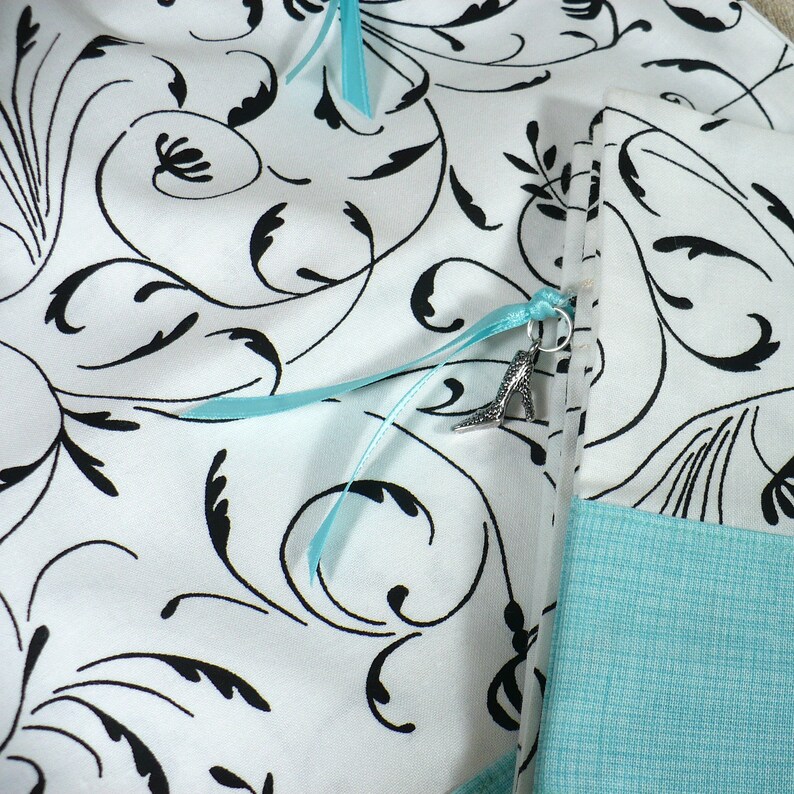 May include: A white fabric pouch with a black floral pattern and a light blue ribbon with a silver shoe charm. The pouch has a light blue fabric lining.