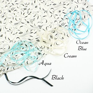 May include: A white fabric bag with a black floral pattern. The bag is decorated with four different colored ribbons: aqua, ocean blue, cream, and black.