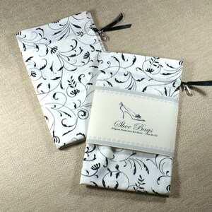 May include: Two white fabric shoe bags with black floral patterns. The bags have black drawstrings with silver beads and a small tag that reads "Shoe Bags Elegant Protection for Shoes on the Go."