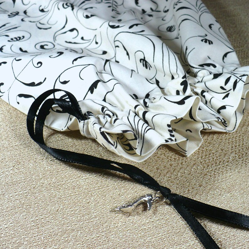 May include: A white fabric drawstring bag with a black and white floral pattern. The bag has a black ribbon drawstring and a silver shoe charm.