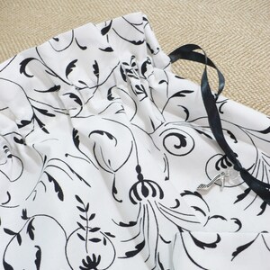 May include: White drawstring bag with a black floral pattern and black ribbon ties. A silver charm is attached to the ribbon.