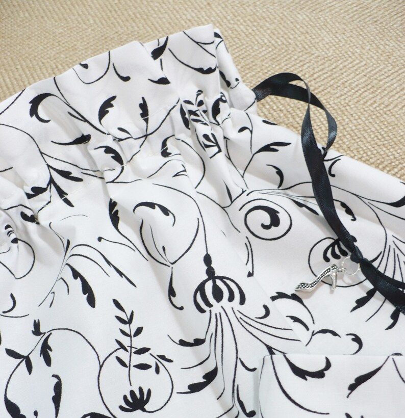 May include: White drawstring bag with a black floral pattern and black ribbon ties. A silver charm is attached to the ribbon.