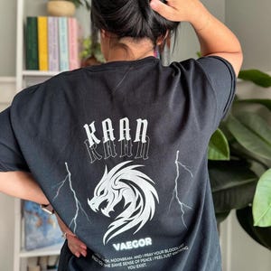 May include: Black t-shirt with a white graphic of a dragon and lightning bolts. The text "KAAN" is printed above the dragon and "VAEGOR" is printed below the dragon. The text "TH MOONBEAM. AND I PRAY YOUR BLOODLUST BRINGS YOU THE SAME SENSE OF PEACE I FEEL JUST KNOWING YOU EXIST." is printed below the barcode.