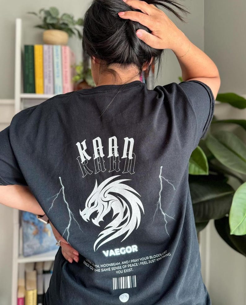 May include: Black t-shirt with a white graphic of a dragon and lightning bolts. The text "KAAN" is printed above the dragon and "VAEGOR" is printed below the dragon. The text "TH MOONBEAM. AND I PRAY YOUR BLOODLUST BRINGS YOU THE SAME SENSE OF PEACE I FEEL JUST KNOWING YOU EXIST." is printed below the barcode.