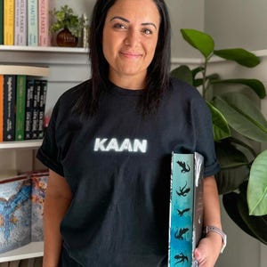 May include: A person wearing a black t-shirt with the word "KAAN" printed on it in white. The person is holding a book with a blue and green gradient cover and black silhouettes of dragons.