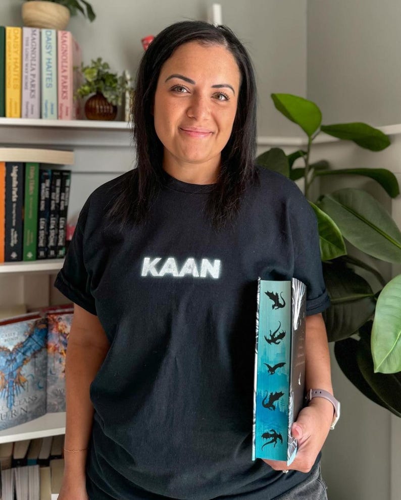 May include: A person wearing a black t-shirt with the word "KAAN" printed on it in white. The person is holding a book with a blue and green gradient cover and black silhouettes of dragons.