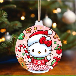 May include: A pink and white Christmas ornament featuring Hello Kitty wearing a red and white Santa hat. The ornament is decorated with candy canes, gingerbread cookies, holly, and a red bow. The text "Name" is printed on the ornament.