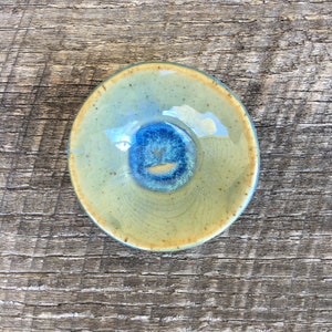 May include: A small, round ceramic dish with a light green glaze and a blue center. The dish has a slightly raised rim and a textured surface.