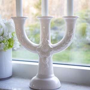 May include: White ceramic candle holder with three arms, each featuring a decorative floral design. The holder is on a windowsill with a bouquet of white flowers in a white vase.