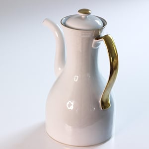 Pode incluir: Cafeteira de cerâmica branca com alça e detalhes dourados. A cafeteira tem uma tampa com botão dourado e um bico fino. O design é simples e elegante, adequado para servir café ou chá.