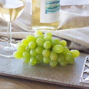 Op de afbeelding: Een stilleven met een glas witte wijn, een fles wijn en een tros groene druiven op een rechthoekig grijs serveerblad. Een decoratief zilverkleurig bladontwerpstuk ligt op de lade.