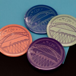 May include: Four round, colorful, pinback buttons with a white outline of a bridge and a train on it. The buttons are blue, green, purple, and orange.
