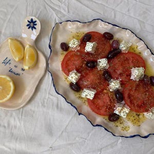Könnte beinhalten: Ein Teller mit geschnittenen roten Tomaten, schwarzen Oliven und weißem Käse, beträufelt mit Öl und Kräutern. Ein kleines Servierbrett mit Zitronenscheiben und dem Text "life is lemon" befindet sich neben dem Teller.
