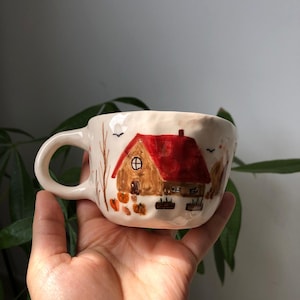 Könnte beinhalten: Eine handbemalte Keramiktasse mit einer herbstlichen Szene. Die Tasse zeigt ein Haus mit rotem Dach, Kürbissen und Bäumen auf cremefarbenem Hintergrund. Sie hat einen gebogenen Henkel und wird in einer Hand gehalten.
