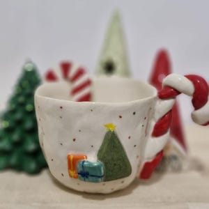 May include: A white ceramic mug with a candy cane handle, decorated with a Christmas tree, presents, and red and white polka dots.