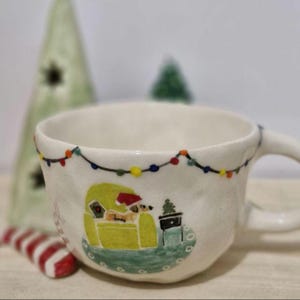 May include: White ceramic mug with a Christmas scene painted on it. The scene features a dog wearing a Santa hat sitting in a green armchair with a Christmas tree next to it. There are colorful lights strung around the top of the mug.