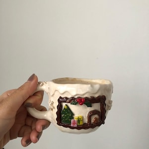 May include: Cream-colored ceramic mug with a decorative Christmas scene. The mug features a brown framed image of a Christmas tree, presents, and a fireplace. The handle and rim have textured details.