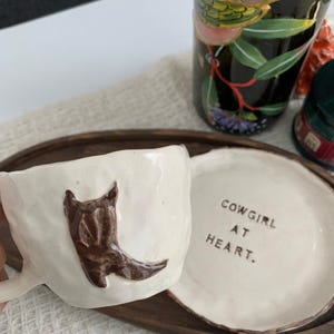 May include: A white ceramic mug with a brown cowboy boot design and a matching saucer. The saucer has the text "COWGIRL AT HEART." The mug and saucer are on a wooden tray, with a decorative bottle in the background.