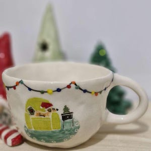 May include: White ceramic mug with a hand-painted design of a dog wearing a Santa hat sitting in a green armchair with a small Christmas tree next to it. The mug has a string of colorful lights around the rim.